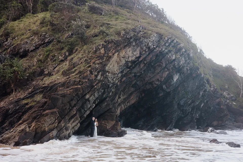 Wide coastal view of Rachel and Dane beneath the sweeping rock formation at Whites Beach. At Whites Beach, Broken Head, near Byron Bay, for an Elopement Collective elopement. Photograph by Jason Lucas.