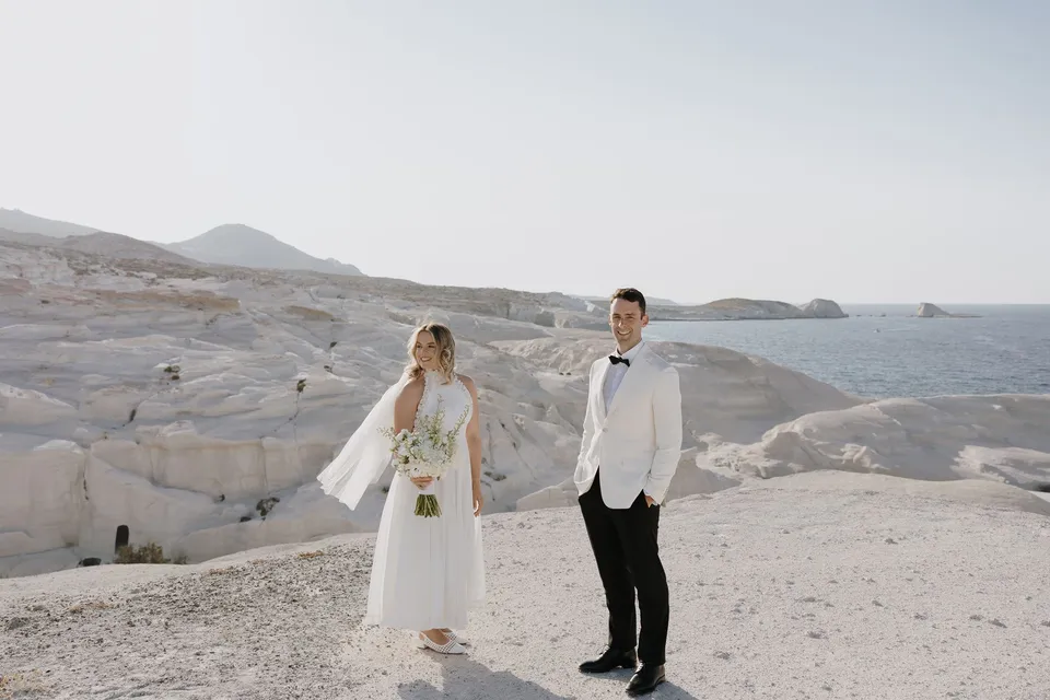 Georgina and Brad facing the camera and holding hands, with her Giasemi bouquet and pale coastal rocks behind them, at Sarakiniko on Milos in the Cyclades, Greece, during an Elopement Collective elopement. Photograph by George Bowden.