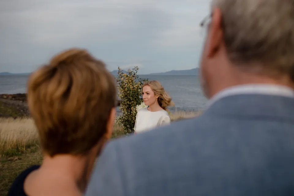 Jemmah listening to Jack’s vows with loved ones in the foreground, at Thalia Haven on the East Coast of Tasmania, Australia, for an Elopement Collective elopement. Photograph by Joey Willis.