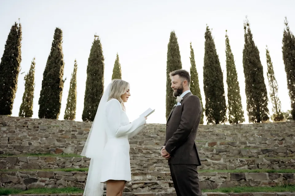 Kaitlyn reading from her vow booklet as Russell holds her hand, with cypress trees behind them. At Piermont Retreat on Tasmania’s east coast, for an Elopement Collective elopement. Photograph by Omar of By The Wilde.