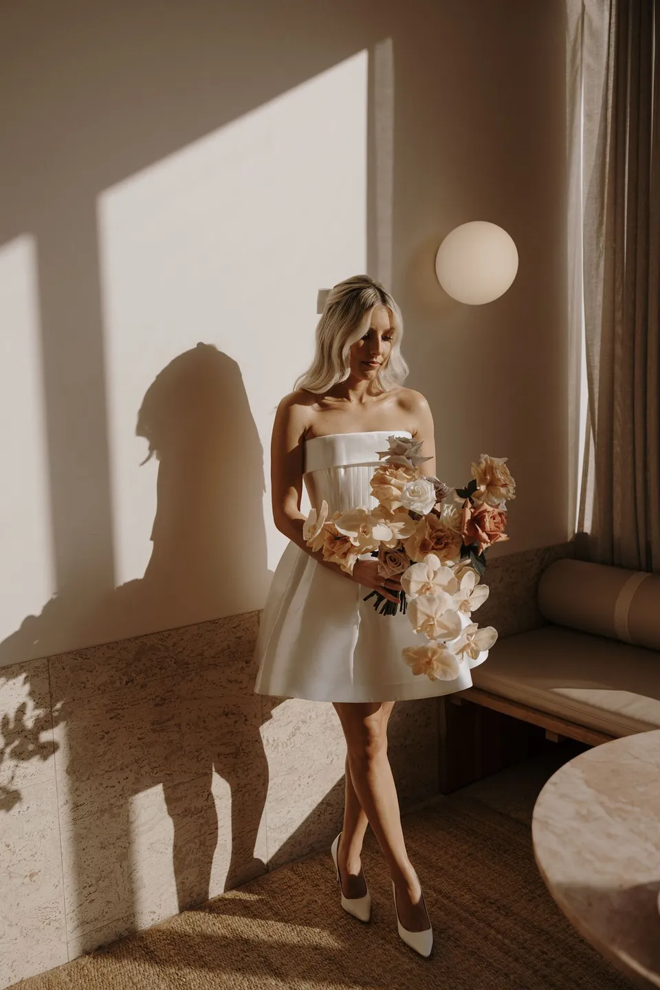 Danielle in her short white dress holding her bouquet beside a sunlit suite window, during Danielle and Adam’s Elopement Collective elopement at The Calile Hotel in Fortitude Valley, Brisbane.