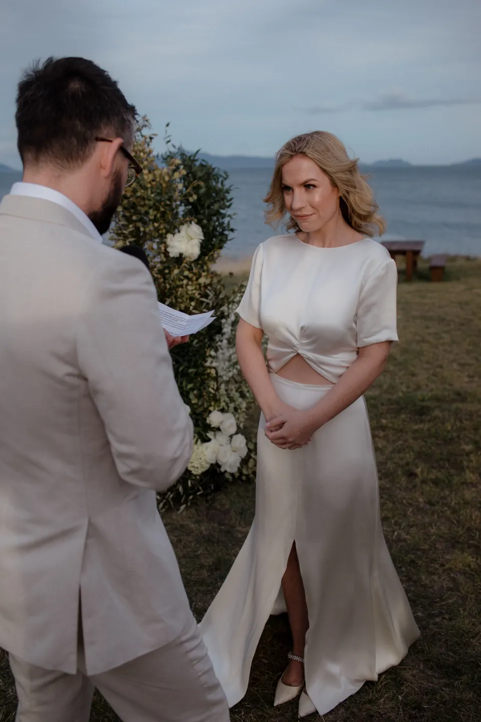 Jemmah listening to Jack beside a ceremony flower arrangement by The Romantics, at Thalia Haven on the East Coast of Tasmania, Australia, for an Elopement Collective elopement. Photograph by Joey Willis.