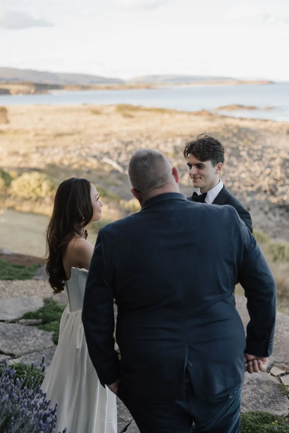 Celebrant Josh Withers speaking with Clara and Harry during their intimate ceremony, with the coast beyond, at Thalia Haven on the East Coast of Tasmania, for an Elopement Collective elopement. Photograph by Bulb Creative.