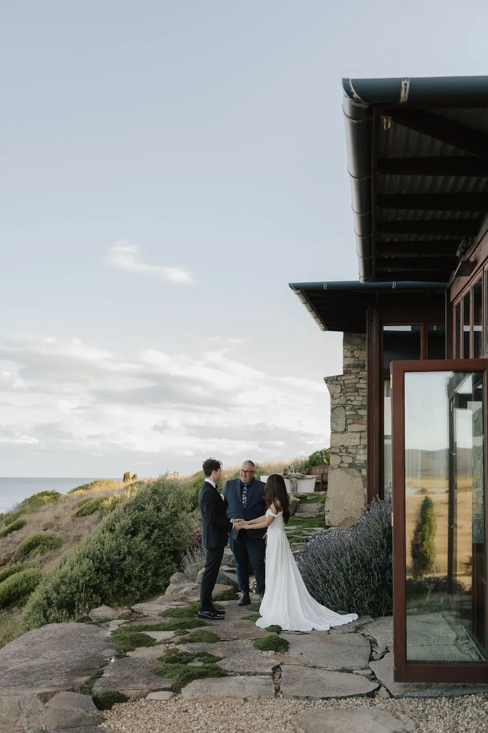 Clara and Harry with celebrant Josh Withers on the stone terrace beside the house, overlooking the sea, at Thalia Haven on the East Coast of Tasmania, for an Elopement Collective elopement. Photograph by Bulb Creative.