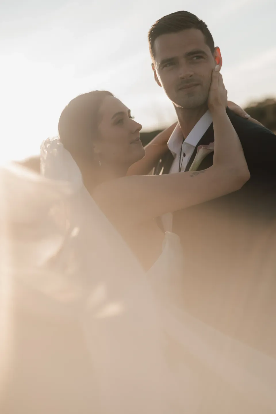 Dani leaning into Anthony with her arms around his neck, photographed through soft golden light, at Thalia Haven on Tasmania’s east coast, for an Elopement Collective elopement. Photograph by Bulb Creative.