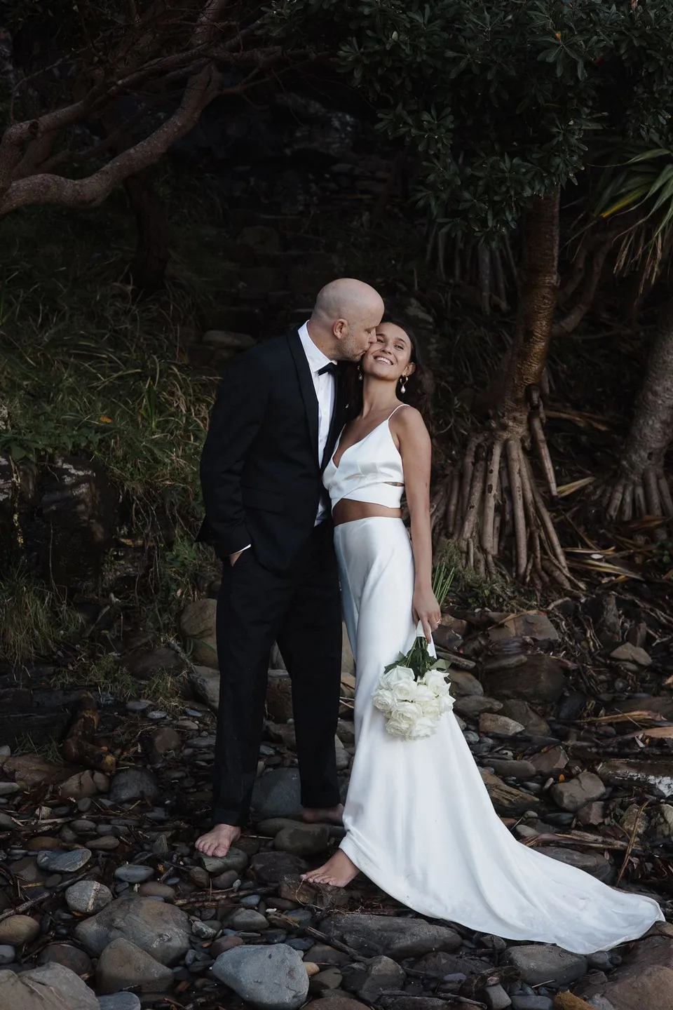 Rachel and Dane embracing beneath a tree beside the beach, her bouquet held at her side, with flowers by Fifth Avenue Florist. At Whites Beach, Broken Head, near Byron Bay, for an Elopement Collective elopement. Photograph by Jason Lucas.