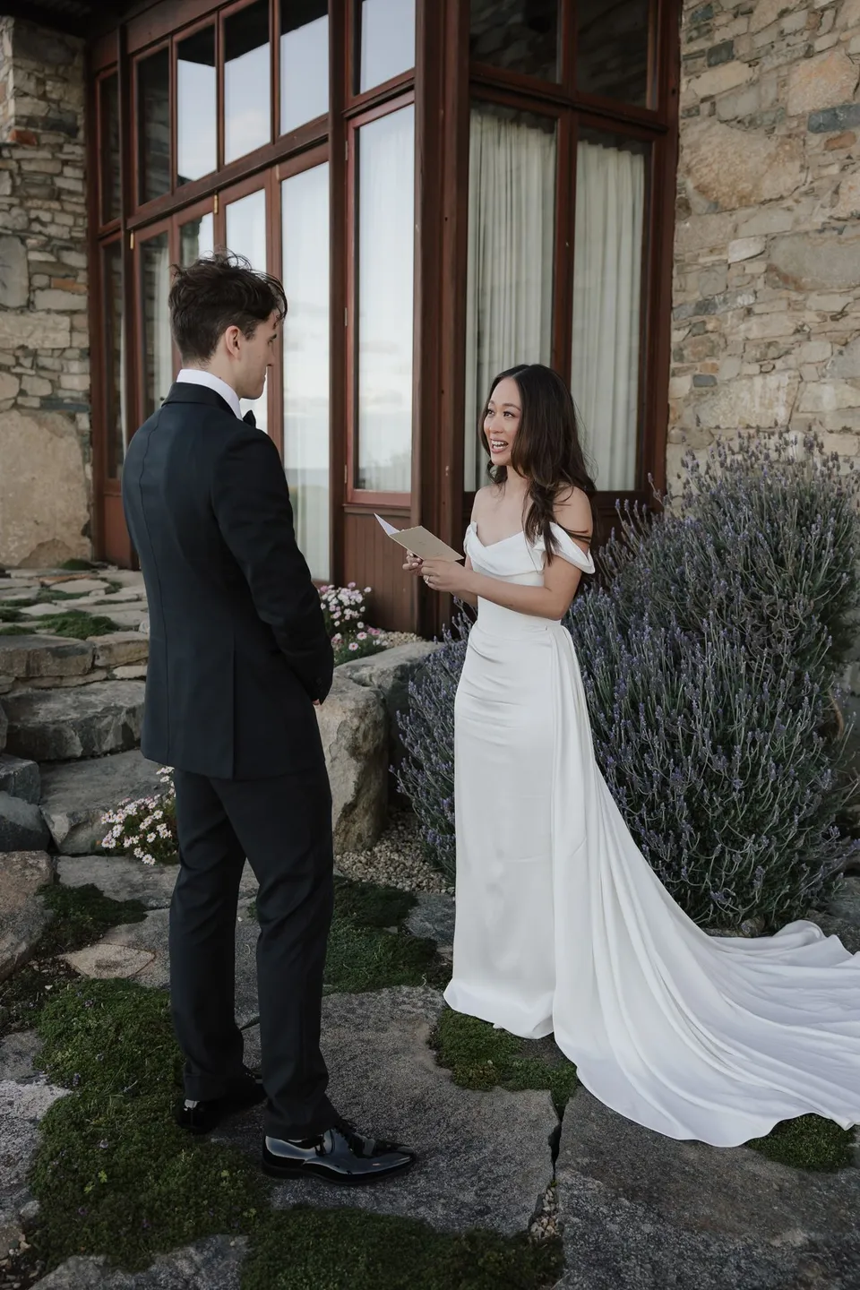 Clara smiling as she reads her personal vows to Harry from an open card beside the stone house and flowering garden, at Thalia Haven on the East Coast of Tasmania, for an Elopement Collective elopement. Photograph by Bulb Creative.