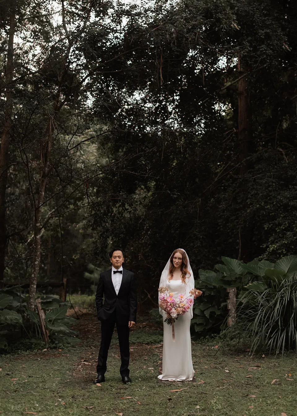 Elyse and Jonathan standing together under the garden canopy with her bouquet, with flowers by The Perfect Party Co. At Fig Tree Villa in Ewingsdale, Byron Bay, for an Elopement Collective elopement. Photograph by Bulb Creative.