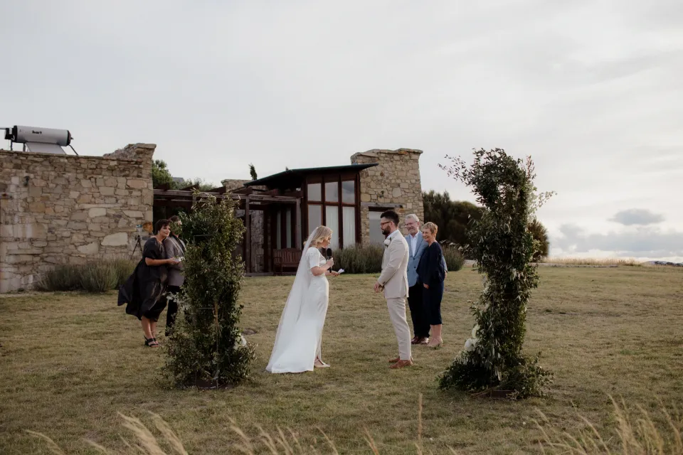Jemmah and Jack exchanging vows on the grass beside the stone buildings, at Thalia Haven on the East Coast of Tasmania, Australia, for an Elopement Collective elopement. Photograph by Joey Willis.
