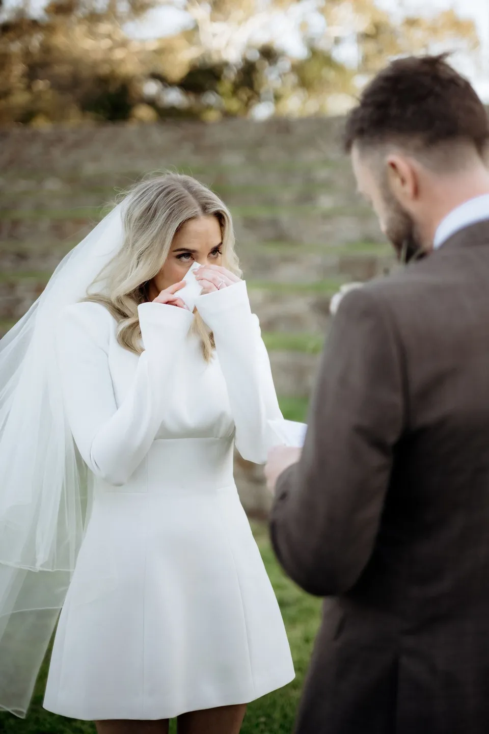 Kaitlyn lifting a tissue to her face while looking at Russell during their vows. At Piermont Retreat on Tasmania’s east coast, for an Elopement Collective elopement. Photograph by Omar of By The Wilde.