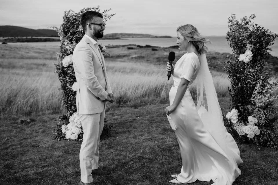 Black and white photograph of Jemmah speaking into the microphone as Jack listens, at Thalia Haven on the East Coast of Tasmania, Australia, for an Elopement Collective elopement. Photograph by Joey Willis.