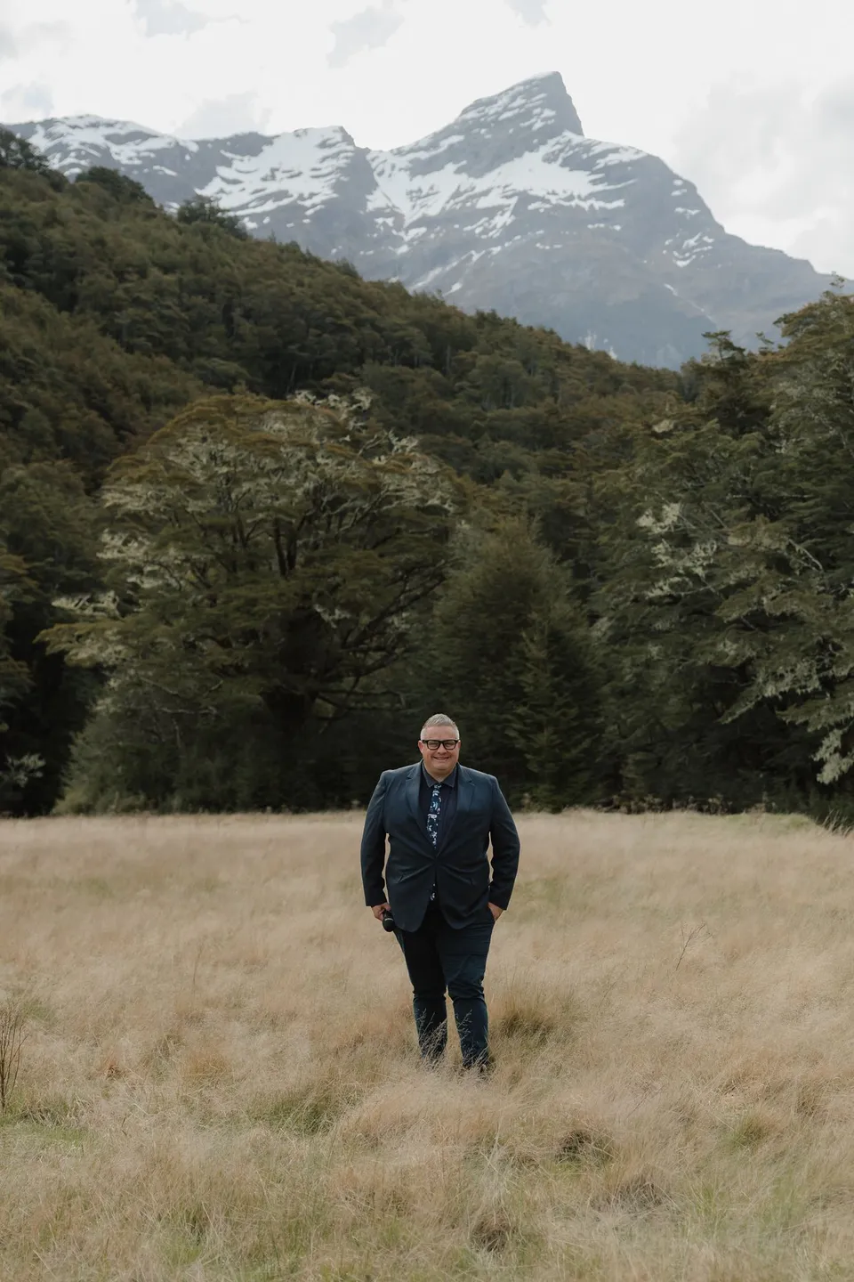 Celebrant Josh Withers smiling with a microphone in the meadow beneath forested slopes and snow-streaked peaks, in the field on the road to Paradise near Glenorchy, in the Queenstown region of New Zealand, for an Elopement Collective Queenstown elopement. Photograph by Bulb Creative.