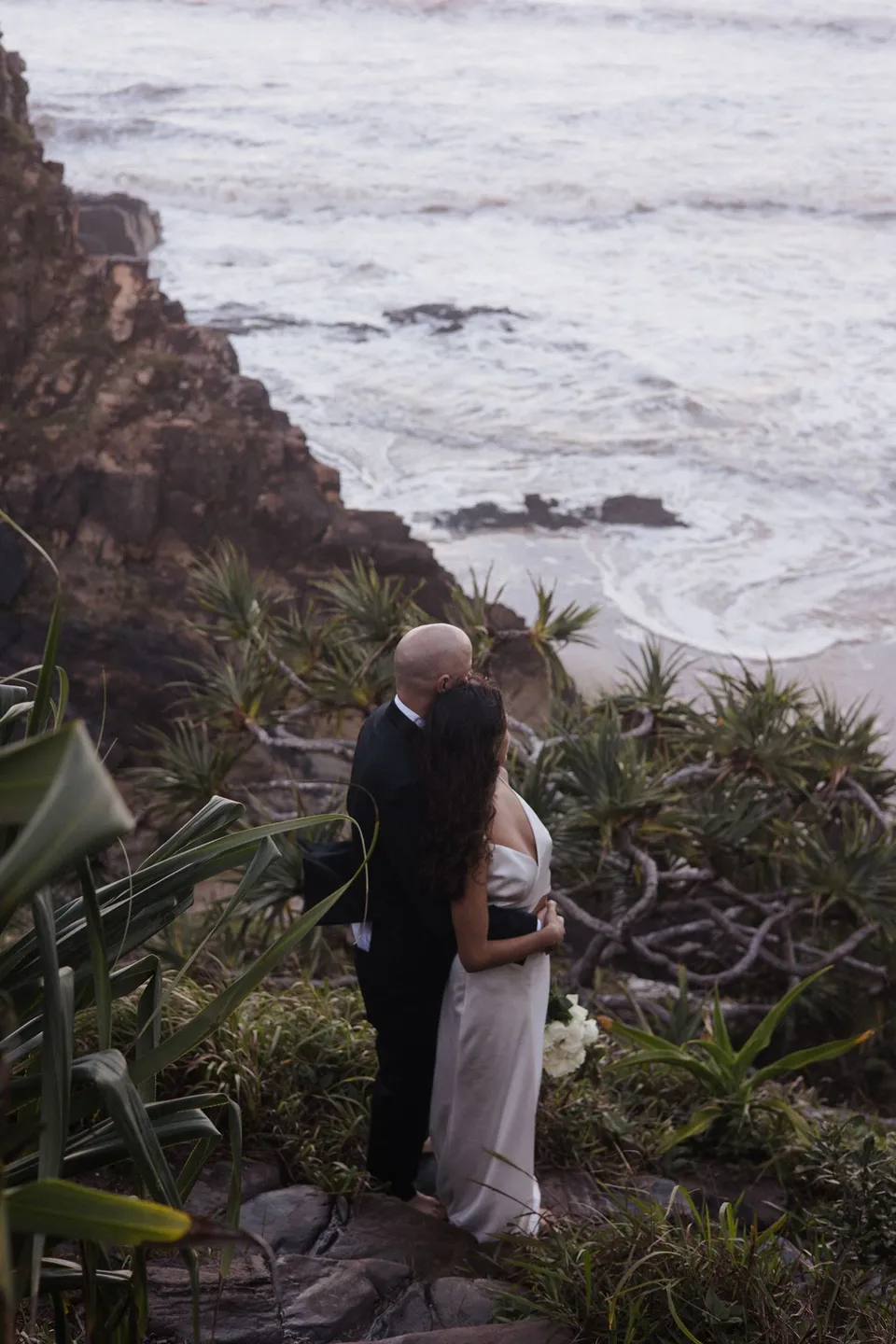 Rachel and Dane embracing beside coastal plants with the ocean below them. At Whites Beach, Broken Head, near Byron Bay, for an Elopement Collective elopement. Photograph by Jason Lucas.