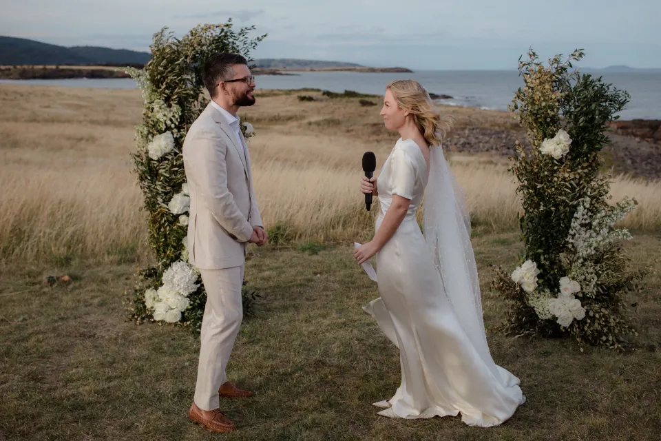 Jemmah reading her personal vows with Jack and the bay behind them, at Thalia Haven on the East Coast of Tasmania, Australia, for an Elopement Collective elopement. Photograph by Joey Willis.