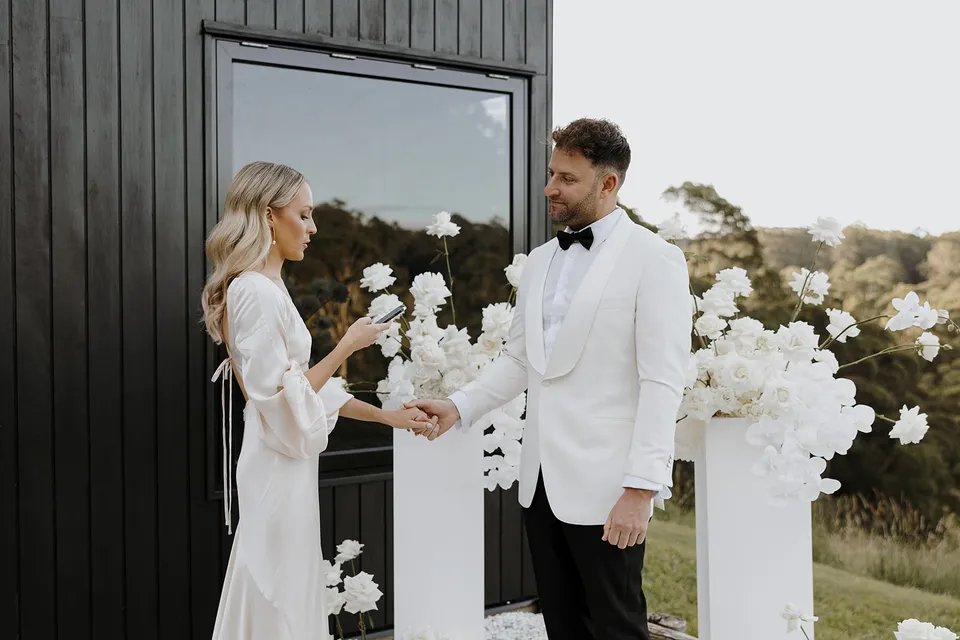 Gabrielle reading her vows while holding Greg’s hand beside the cabin’s dark timber wall at Roundhill Retreat in Obi Obi, Sunshine Coast Hinterland, for their Elopement Collective elopement.