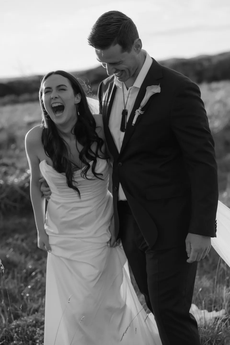 Black and white portrait of Dani laughing beside Anthony in the coastal grassland, at Thalia Haven on Tasmania’s east coast, for an Elopement Collective elopement. Photograph by Bulb Creative.