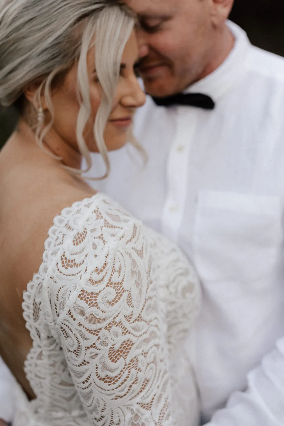 A close portrait of Matt embracing Laura, showing her lace sleeves and low-backed dress, in the Tweed Valley, NSW, for an Elopement Collective elopement. Photo by Bulb Creative.