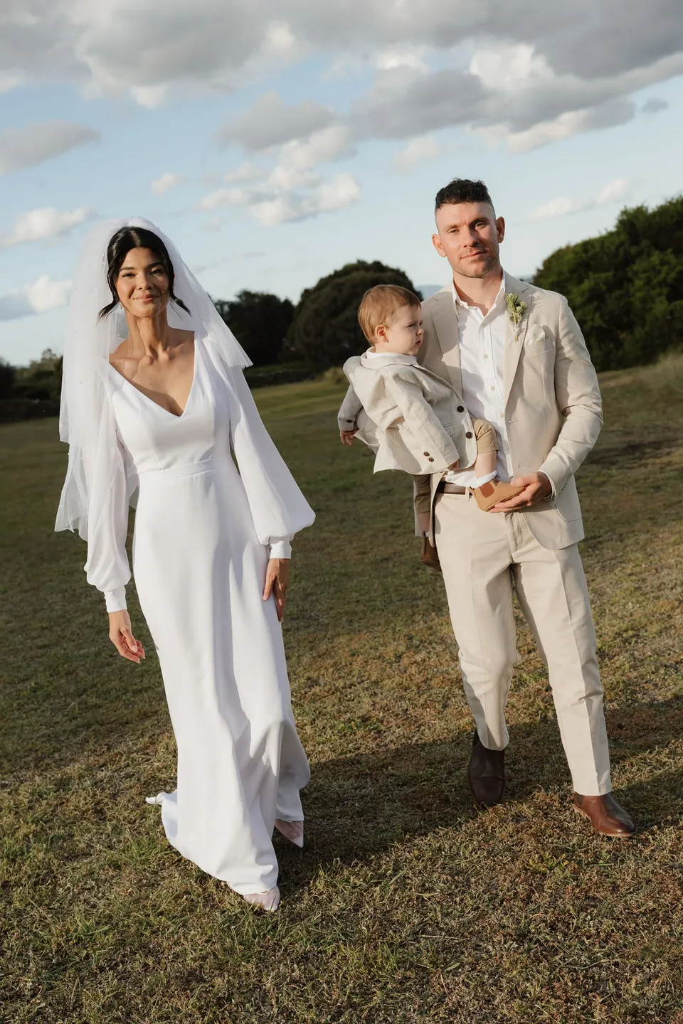 Renee and Mitch standing together on the lawn after their ceremony, with Mitch holding a young child. At Piermont Retreat on Tasmania’s east coast, for an Elopement Collective elopement. Photograph by Bulb Creative.