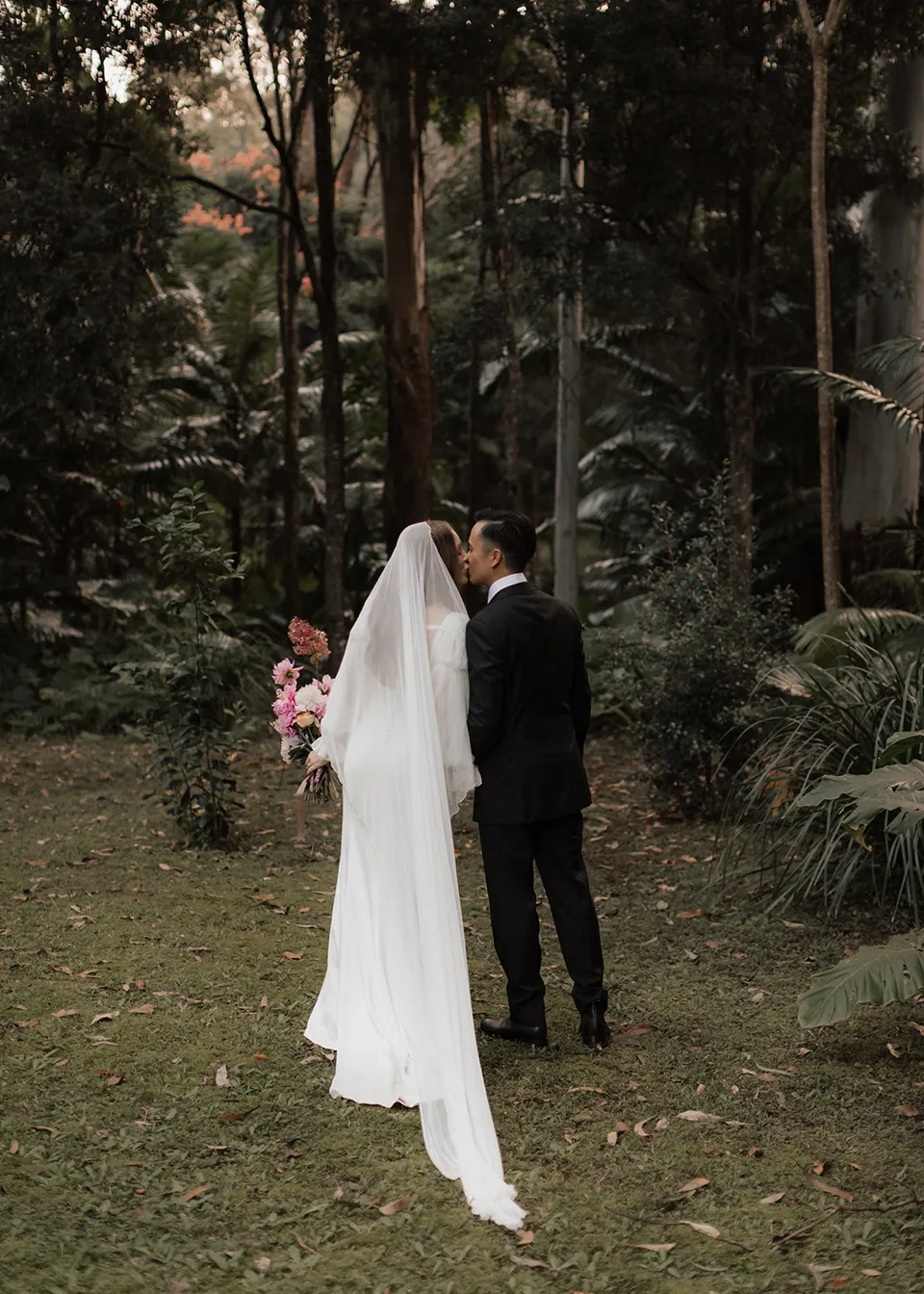 Elyse and Jonathan pausing together among the trees with her veil trailing behind them. At Fig Tree Villa in Ewingsdale, Byron Bay, for an Elopement Collective elopement. Photograph by Bulb Creative.