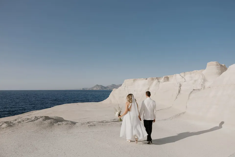 Georgina and Brad walking beneath white cliffs beside blue water, casting long shadows across the rocks, at Sarakiniko on Milos in the Cyclades, Greece, during an Elopement Collective elopement. Photograph by George Bowden.