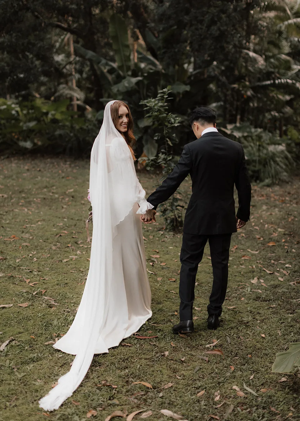 Elyse smiling back over her shoulder as Jonathan leads her through the garden. At Fig Tree Villa in Ewingsdale, Byron Bay, for an Elopement Collective elopement. Photograph by Bulb Creative.