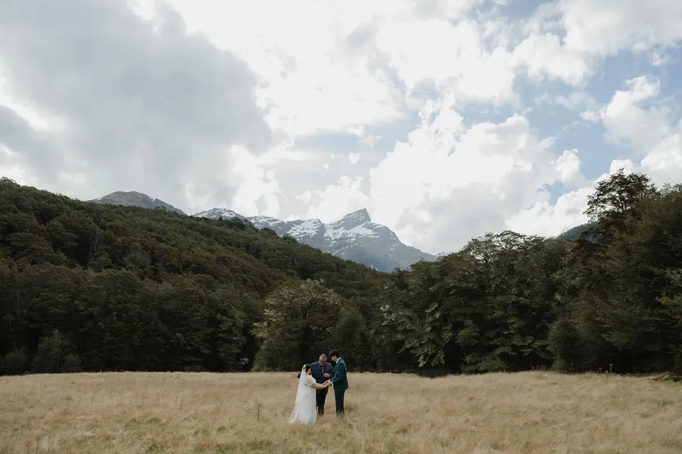Jaeda and Adrian with celebrant Josh Withers in a wide grassy field surrounded by forest and snow-streaked mountains, in the field on the road to Paradise near Glenorchy, in the Queenstown region of New Zealand, for an Elopement Collective Queenstown elopement. Photograph by Bulb Creative.