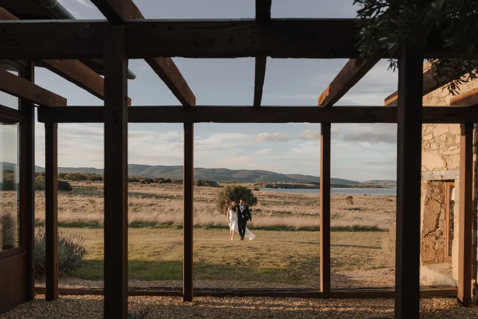 Dani and Anthony walking towards a timber-framed stone courtyard, with golden grassland and Great Oyster Bay beyond, at Thalia Haven on Tasmania’s east coast, for an Elopement Collective elopement. Photograph by Bulb Creative.
