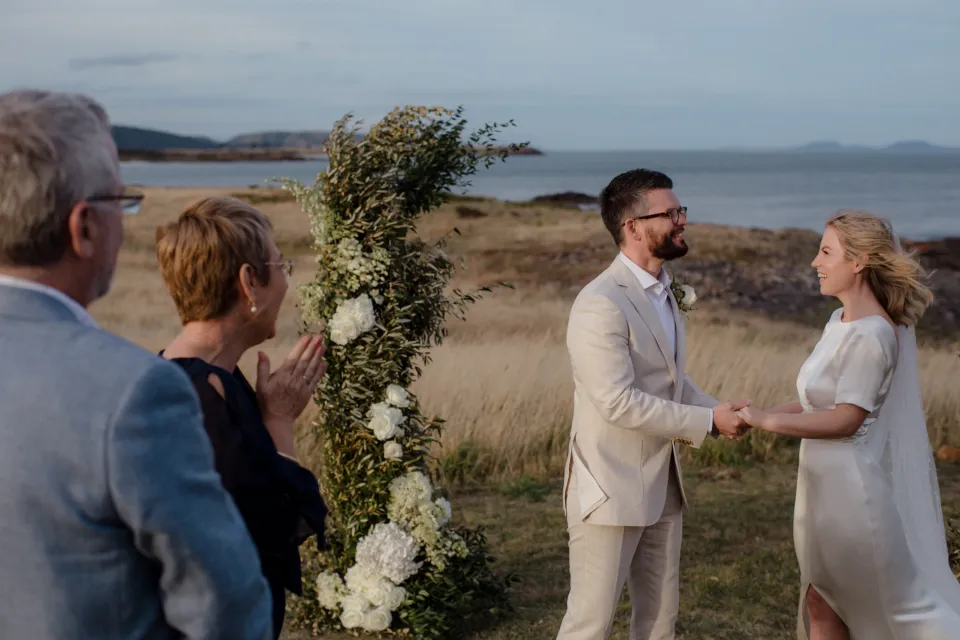 Jemmah and Jack holding hands and smiling with guests nearby, at Thalia Haven on the East Coast of Tasmania, Australia, for an Elopement Collective elopement. Photograph by Joey Willis.