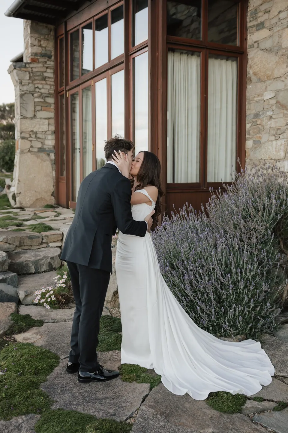 Clara and Harry kissing after their ceremony beside the stone house, with her dress train across the paving, at Thalia Haven on the East Coast of Tasmania, for an Elopement Collective elopement. Photograph by Bulb Creative.