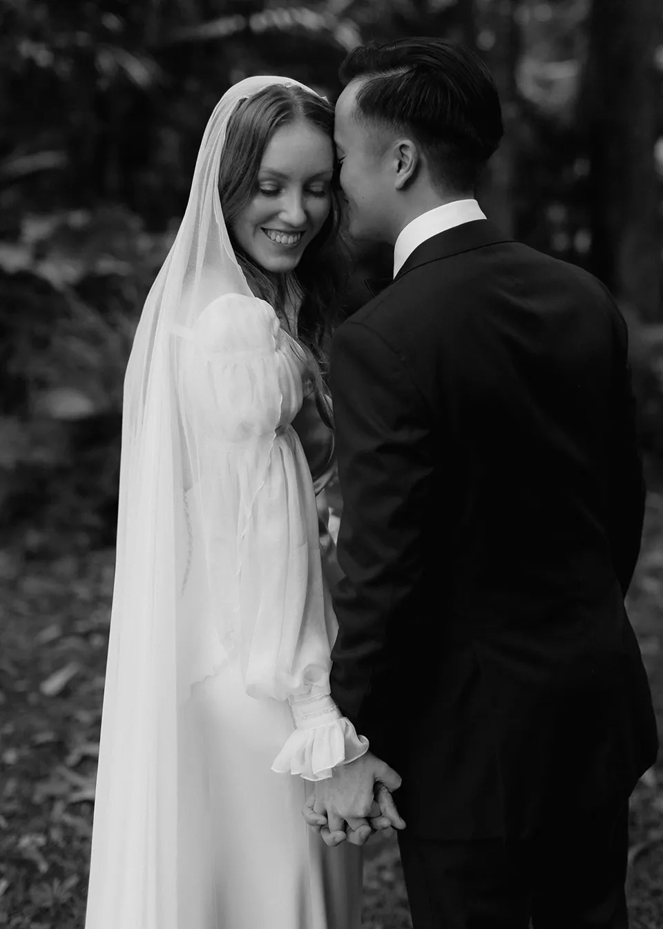 Black-and-white portrait of Elyse smiling beside Jonathan among the trees. At Fig Tree Villa in Ewingsdale, Byron Bay, for an Elopement Collective elopement. Photograph by Bulb Creative.