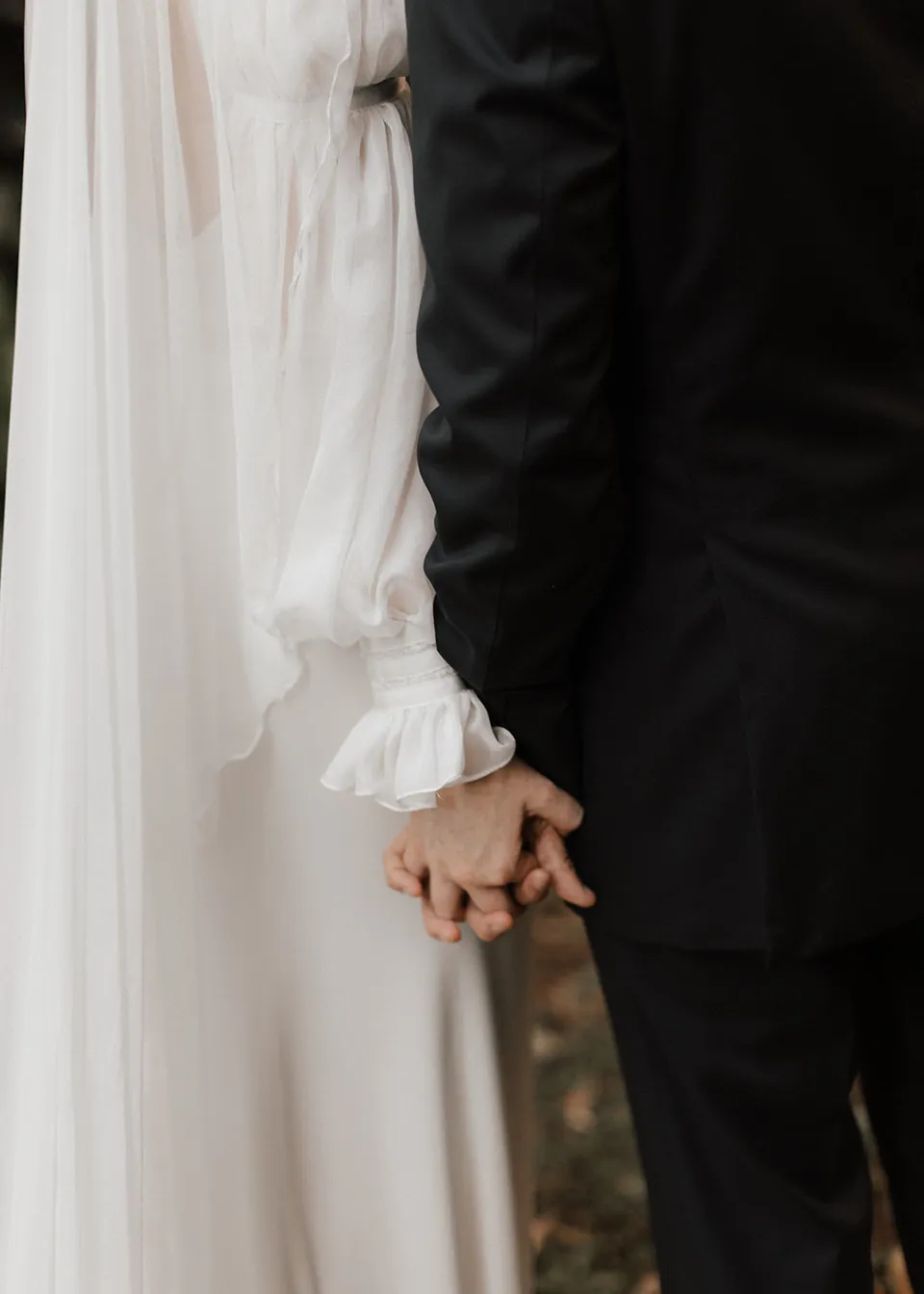 Close-up of the couple's joined hands beside Elyse's white dress. At Fig Tree Villa in Ewingsdale, Byron Bay, for an Elopement Collective elopement. Photograph by Bulb Creative.