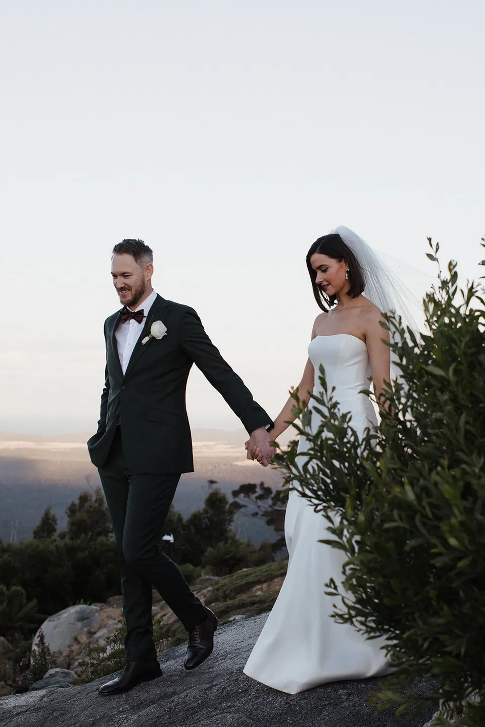 Meg and Ryan smiling as they walk hand in hand across the rocks. At The Keep on Tasmania’s east coast, for an Elopement Collective elopement. Photograph by Jenna Agius.