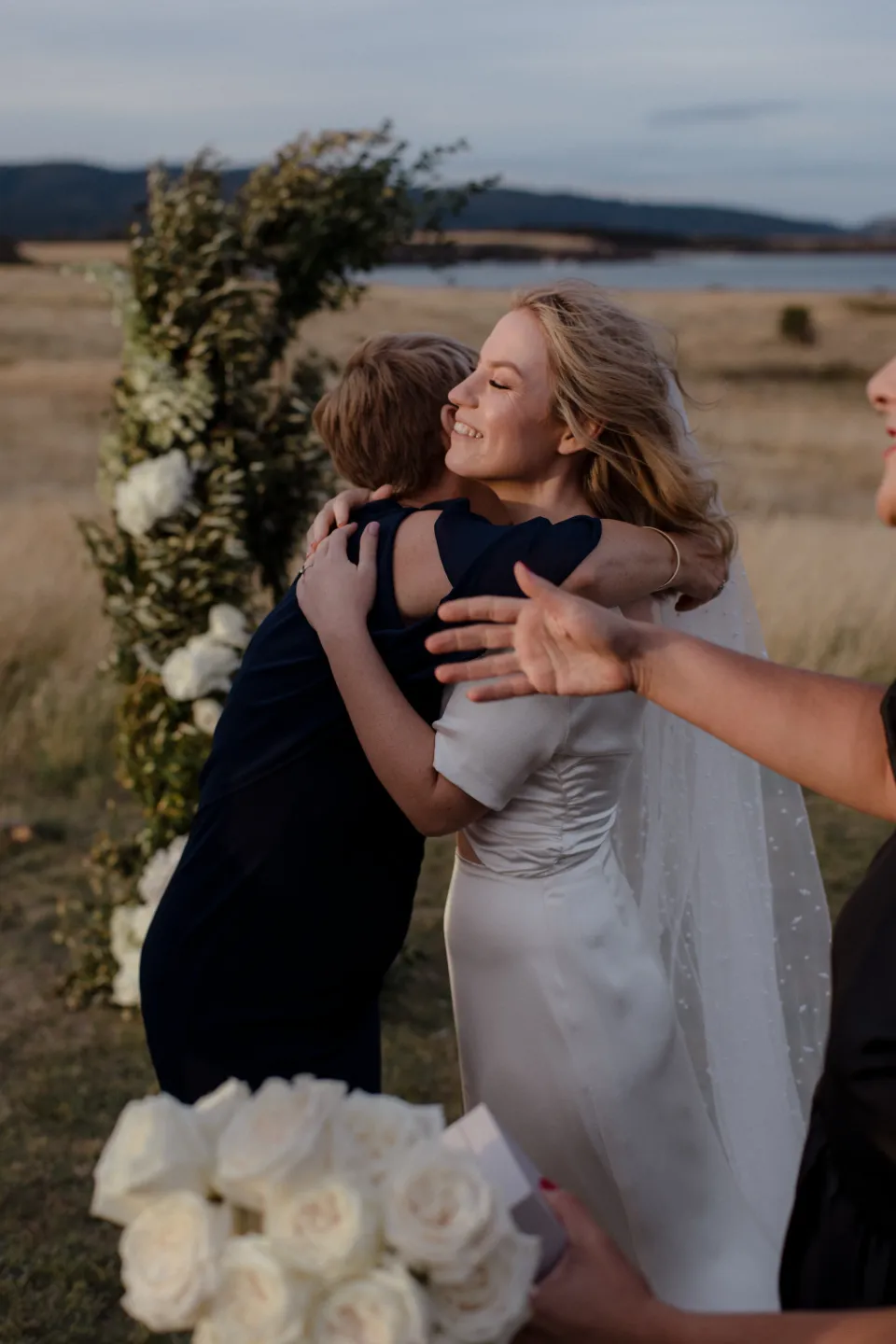 Jemmah smiling as she embraces a loved one beside the ceremony flowers, at Thalia Haven on the East Coast of Tasmania, Australia, for an Elopement Collective elopement. Photograph by Joey Willis.