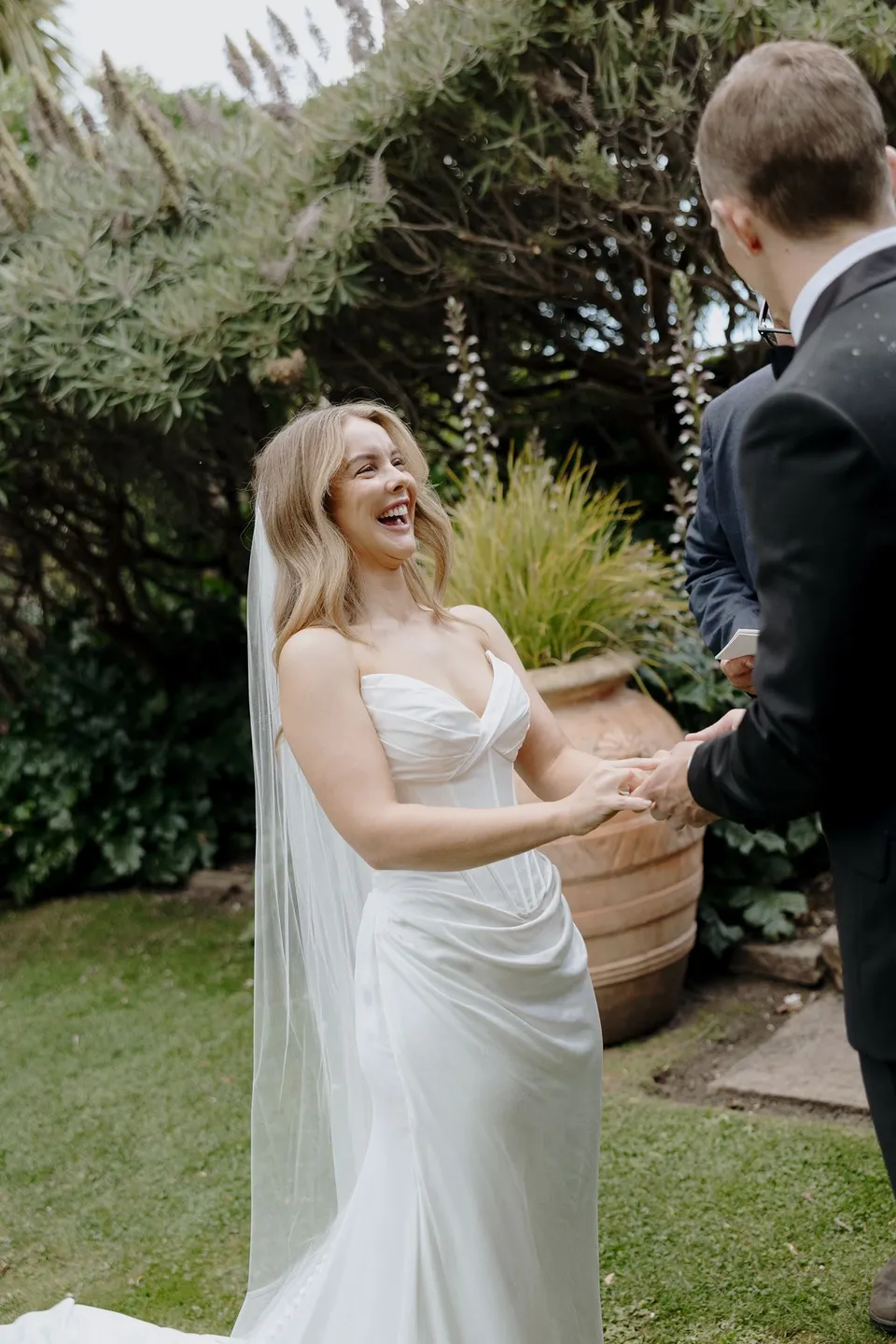Tanesha and Andrew laughing together while holding hands in the leafy hotel garden, at Islington Hotel in South Hobart, Tasmania, Australia, for an Elopement Collective summer elopement. Photograph by George Bowden.