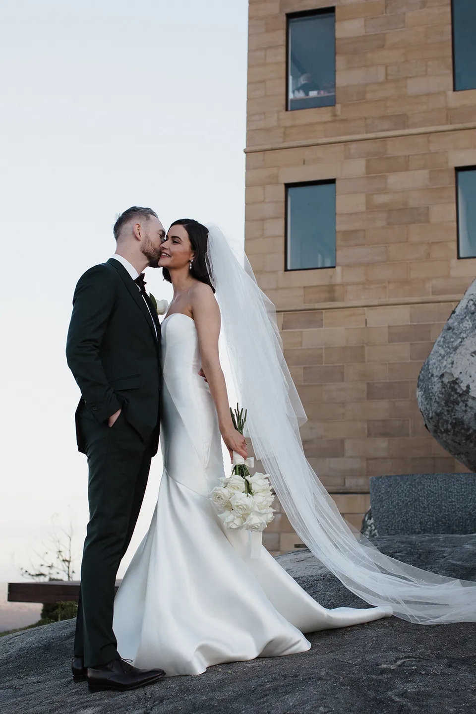 Meg and Ryan kissing beside the sandstone tower, with her long veil trailing behind her. At The Keep on Tasmania’s east coast, for an Elopement Collective elopement. Photograph by Jenna Agius.