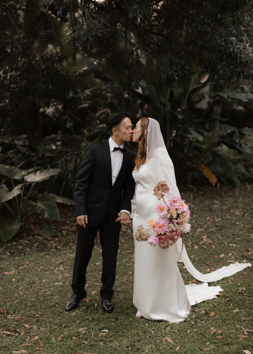The couple sharing a kiss in the garden with flowers at Elyse's side. At Fig Tree Villa in Ewingsdale, Byron Bay, for an Elopement Collective elopement. Photograph by Bulb Creative.