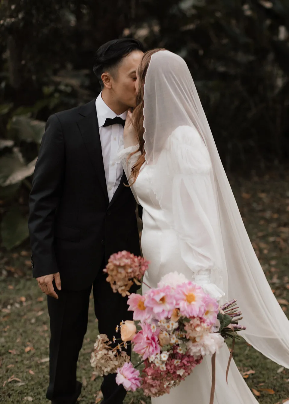 Jonathan kissing Elyse beneath her veil beside the pink bouquet, with flowers by The Perfect Party Co. At Fig Tree Villa in Ewingsdale, Byron Bay, for an Elopement Collective elopement. Photograph by Bulb Creative.