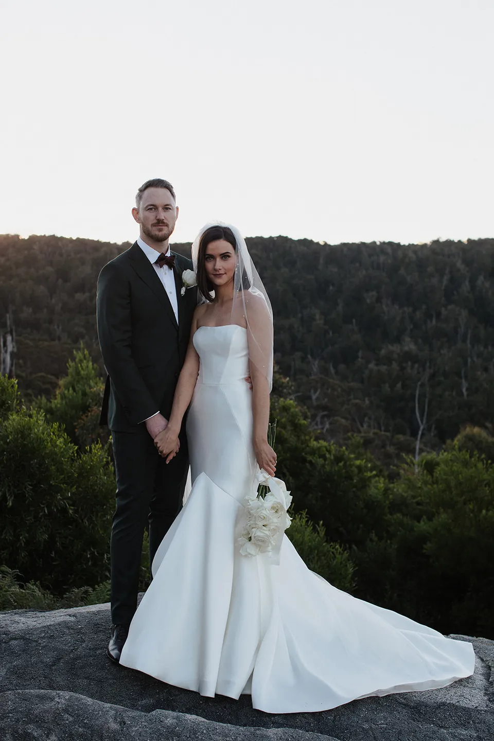 Meg and Ryan standing together on a broad rock, with her dress and veil spread out against the forest view. At The Keep on Tasmania’s east coast, for an Elopement Collective elopement. Photograph by Jenna Agius.