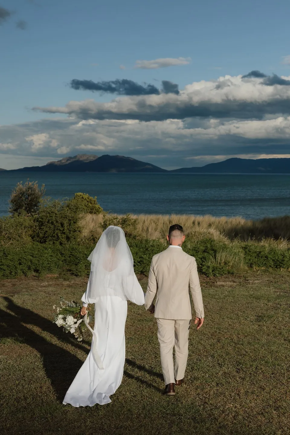 Renee and Mitch walking hand in hand towards the bay, with her veil, dress train and bouquet by The Romantics. At Piermont Retreat on Tasmania’s east coast, for an Elopement Collective elopement. Photograph by Bulb Creative.