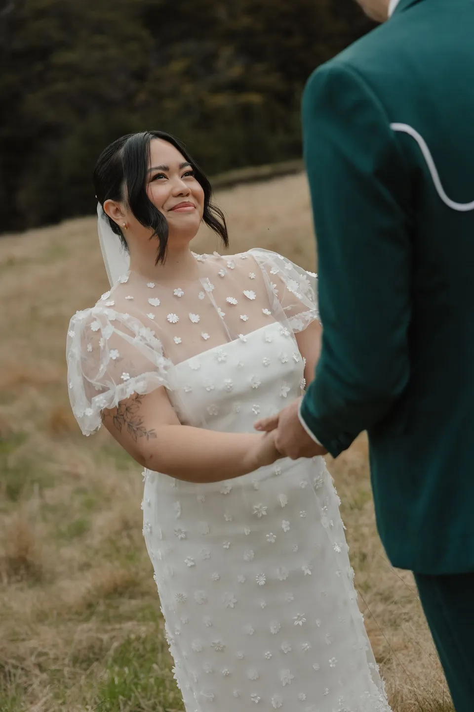 Jaeda smiling up at Adrian as they hold hands during their intimate outdoor ceremony, in the field on the road to Paradise near Glenorchy, in the Queenstown region of New Zealand, for an Elopement Collective Queenstown elopement. Photograph by Bulb Creative.