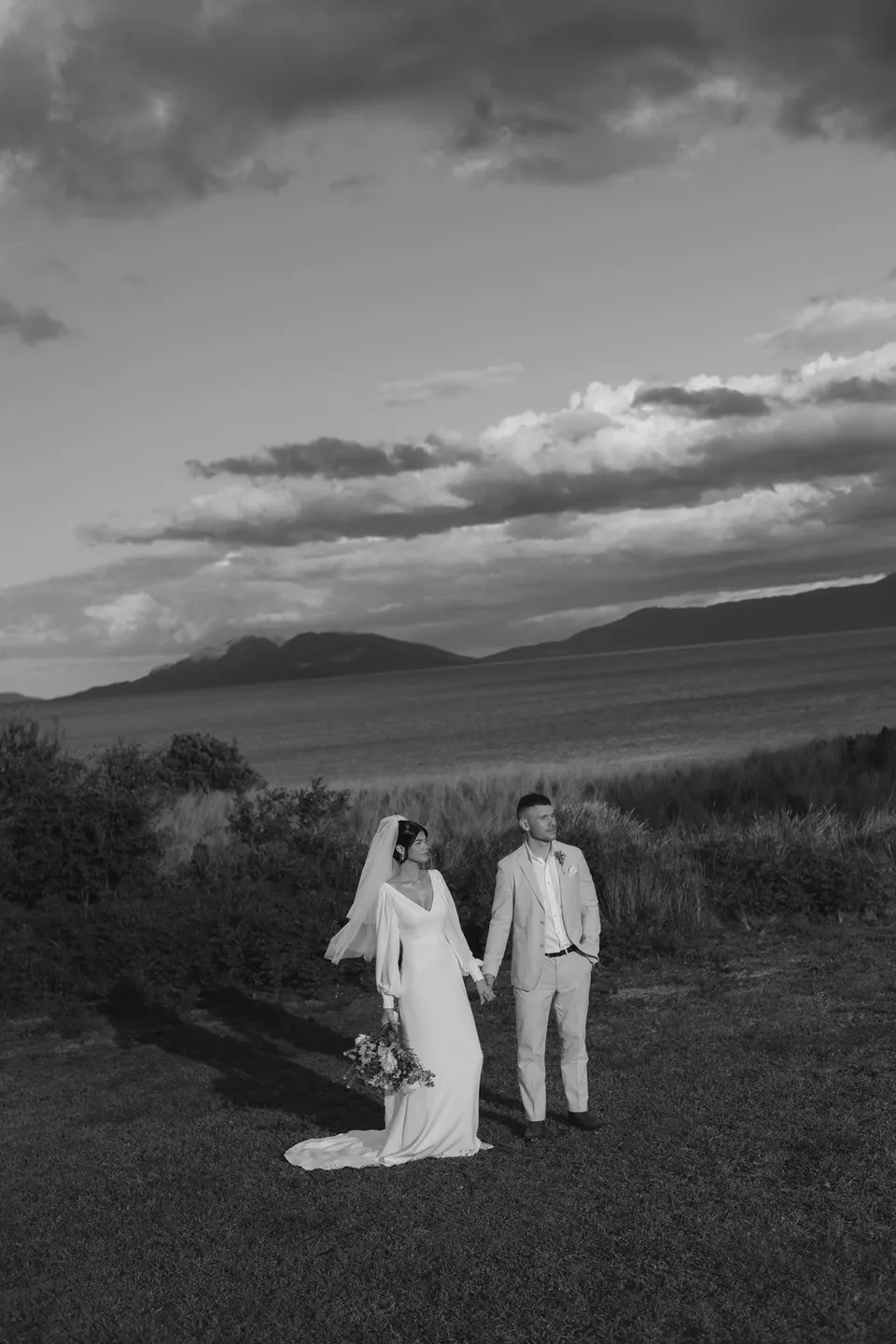 Black and white portrait of Renee and Mitch holding hands on the lawn above the bay, with her bouquet by The Romantics. At Piermont Retreat on Tasmania’s east coast, for an Elopement Collective elopement. Photograph by Bulb Creative.