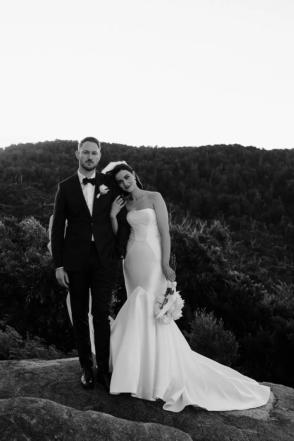 Black and white portrait of Meg leaning towards Ryan on the rocky hilltop, with forested hills behind them. At The Keep on Tasmania’s east coast, for an Elopement Collective elopement. Photograph by Jenna Agius.