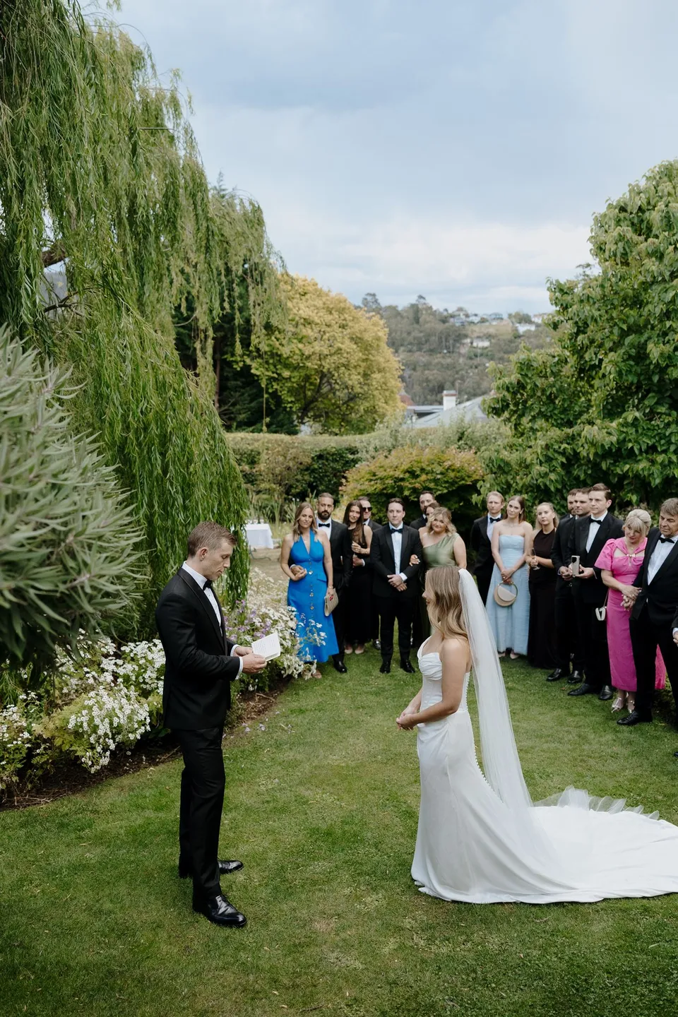 Tanesha and Andrew facing each other on the lawn as their friends and family watch beside a weeping willow, at Islington Hotel in South Hobart, Tasmania, Australia, for an Elopement Collective summer elopement. Photograph by George Bowden.