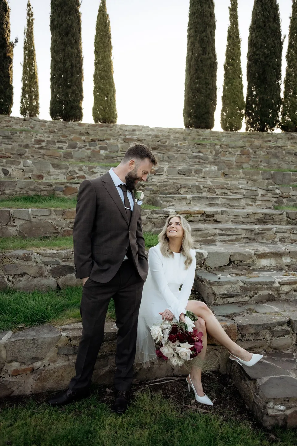 Kaitlyn laughing up at Russell while seated on the stone steps, holding her bouquet by The Romantics. At Piermont Retreat on Tasmania’s east coast, for an Elopement Collective elopement. Photograph by Omar of By The Wilde.