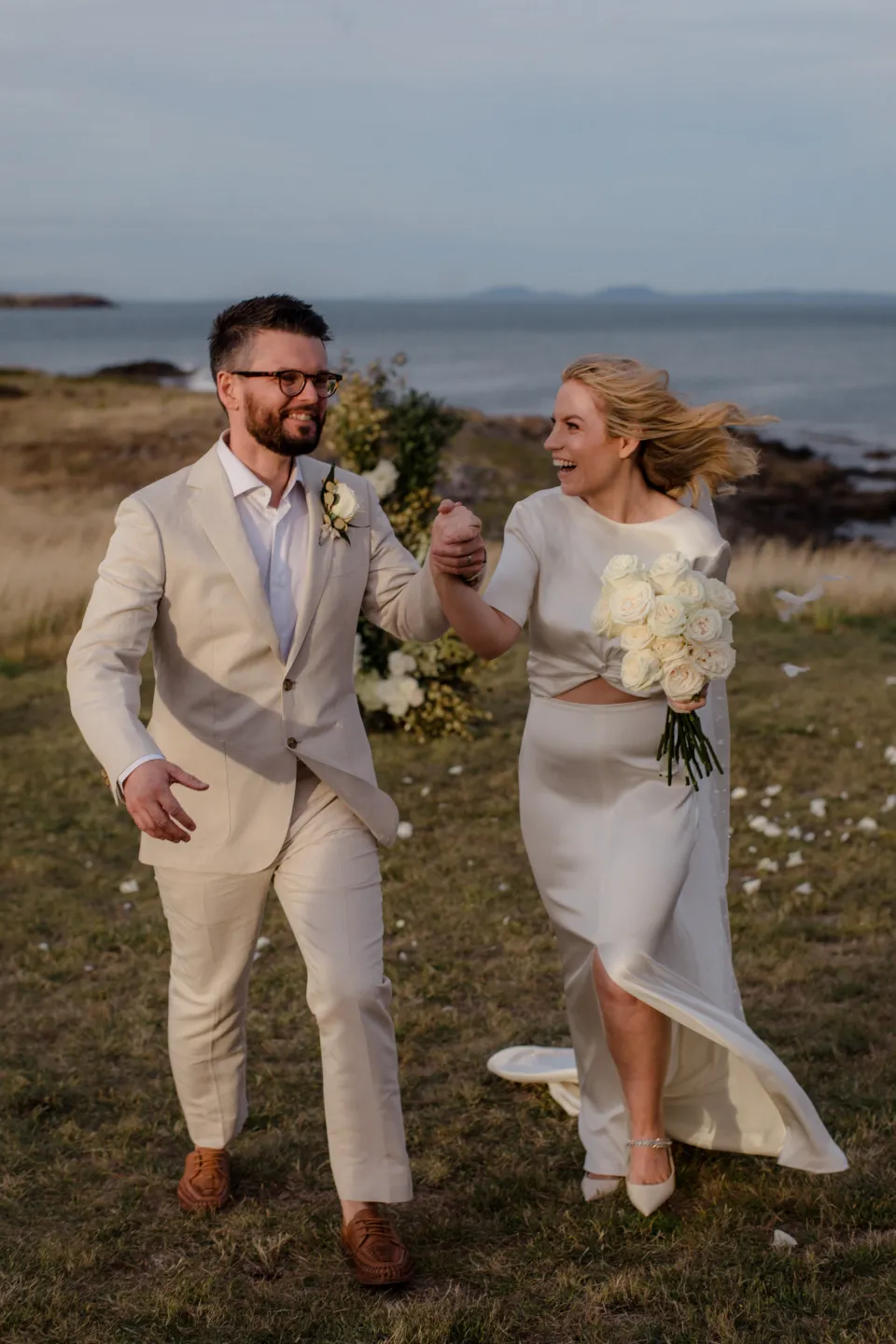 Jemmah and Jack laughing as they walk with her bouquet by The Romantics, at Thalia Haven on the East Coast of Tasmania, Australia, for an Elopement Collective elopement. Photograph by Joey Willis.