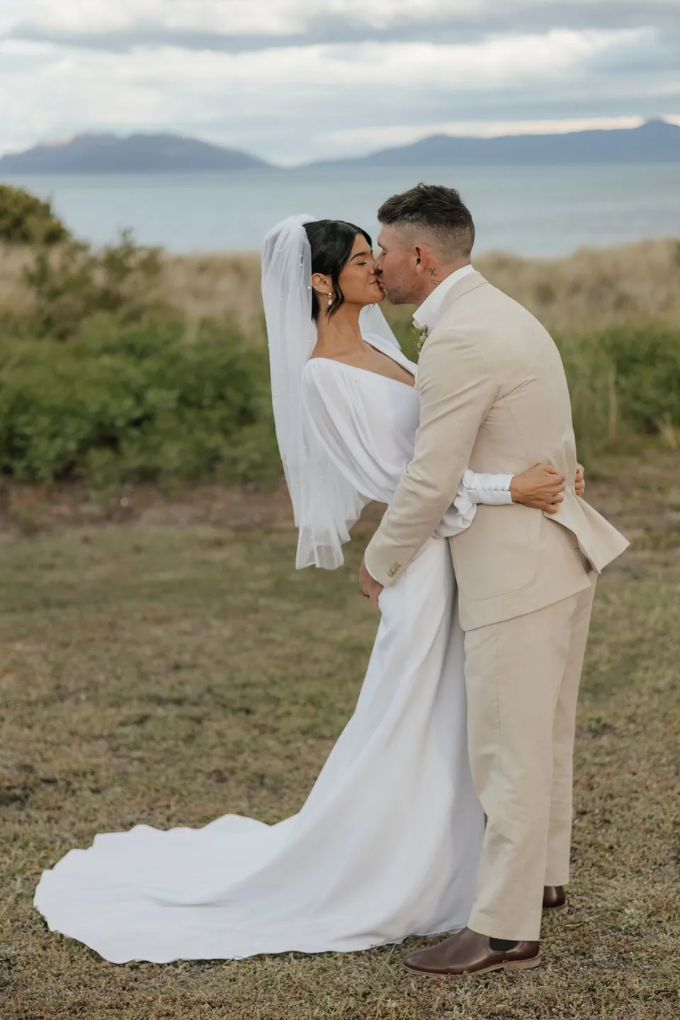 Renee and Mitch kissing on the coastal lawn, with her dress train spread over the grass and distant hills across the water. At Piermont Retreat on Tasmania’s east coast, for an Elopement Collective elopement. Photograph by Bulb Creative.