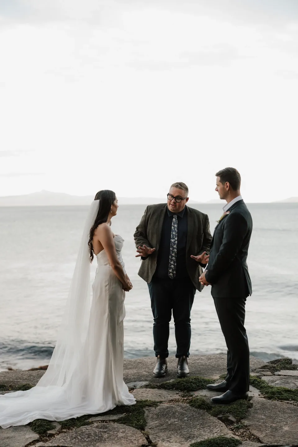 Celebrant Josh Withers speaking with Dani and Anthony during their ceremony beside the still water of Great Oyster Bay, at Thalia Haven on Tasmania’s east coast, for an Elopement Collective elopement. Photograph by Bulb Creative.