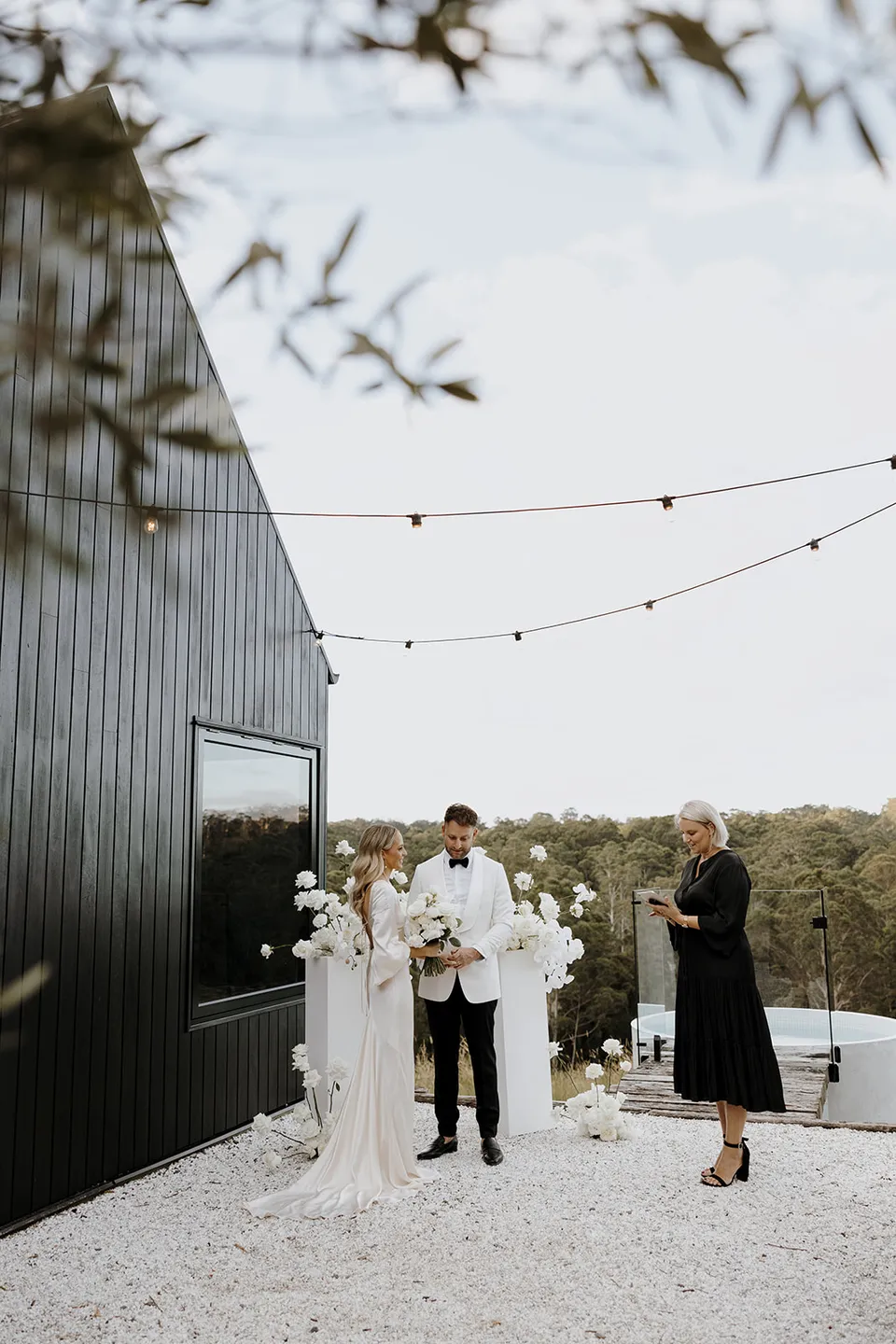 Wide view of Gabrielle and Greg with celebrant Hayley Hart beside the cabin, white flowers and hinterland trees at Roundhill Retreat in Obi Obi, Sunshine Coast Hinterland, for their Elopement Collective elopement.
