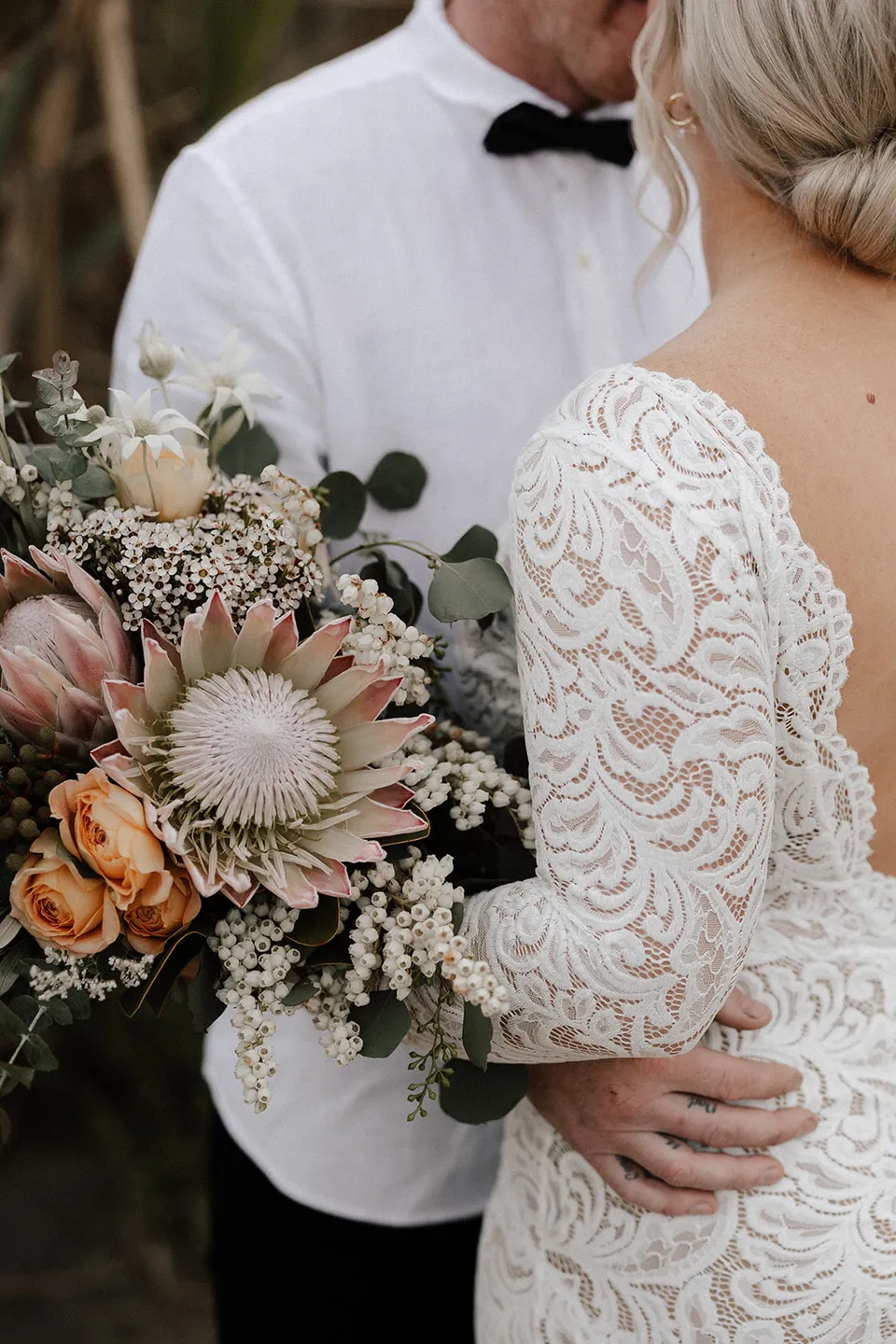 Laura’s bouquet by Fifth Avenue Florist, with proteas, roses and eucalyptus, beside her lace dress and Matt’s white shirt, in the Tweed Valley, NSW, for an Elopement Collective elopement. Photo by Bulb Creative.