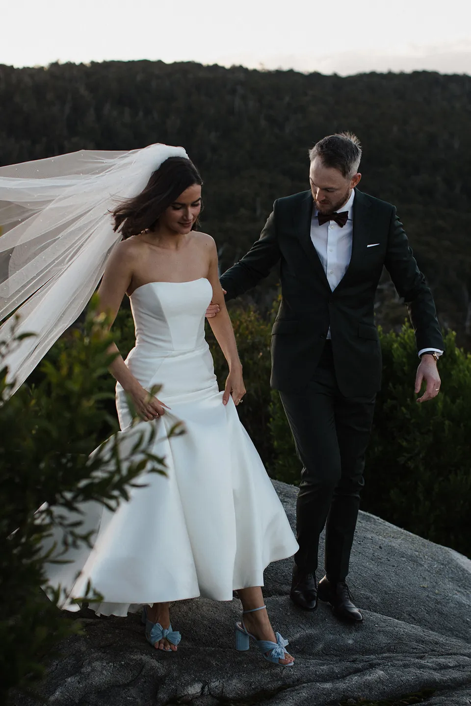 Meg and Ryan carefully stepping down a rock, with her veil lifting in the breeze and blue shoes visible. At The Keep on Tasmania’s east coast, for an Elopement Collective elopement. Photograph by Jenna Agius.